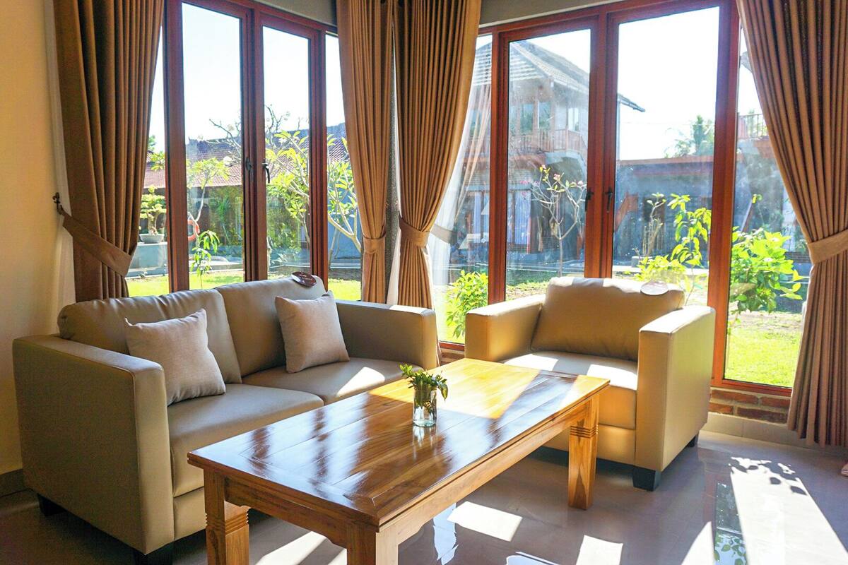A living room with a sofa, a coffee table, and a chair. There are large windows with curtains.