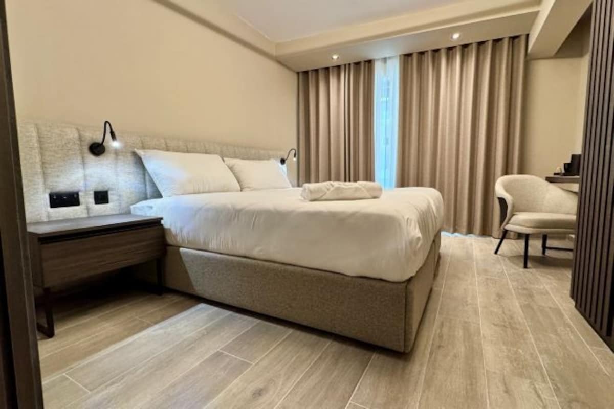 A modern hotel room with a large bed, a bedside table, a chair, and a large window with curtains.