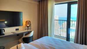 A hotel room with a bed, a desk with a chair, a TV, and a view of the sea through a large window. - Aste Hotel (St. Julian's)