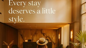 A woman in a dress and hat walking with a suitcase in a hotel lobby with a reception desk and seating area. - Aste Hotel (St. Julian's)