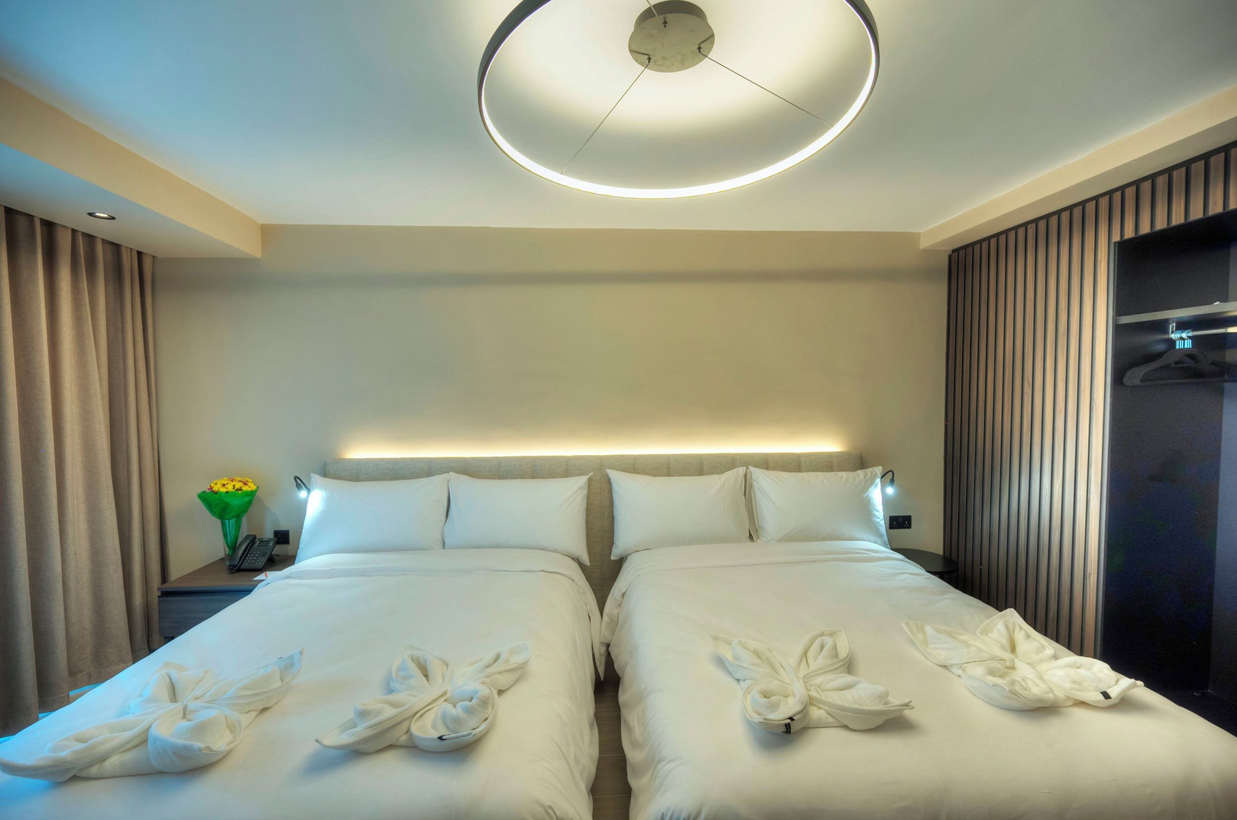 A hotel room with two beds, a round ceiling light, a nightstand with a phone, and a wall-mounted TV.
