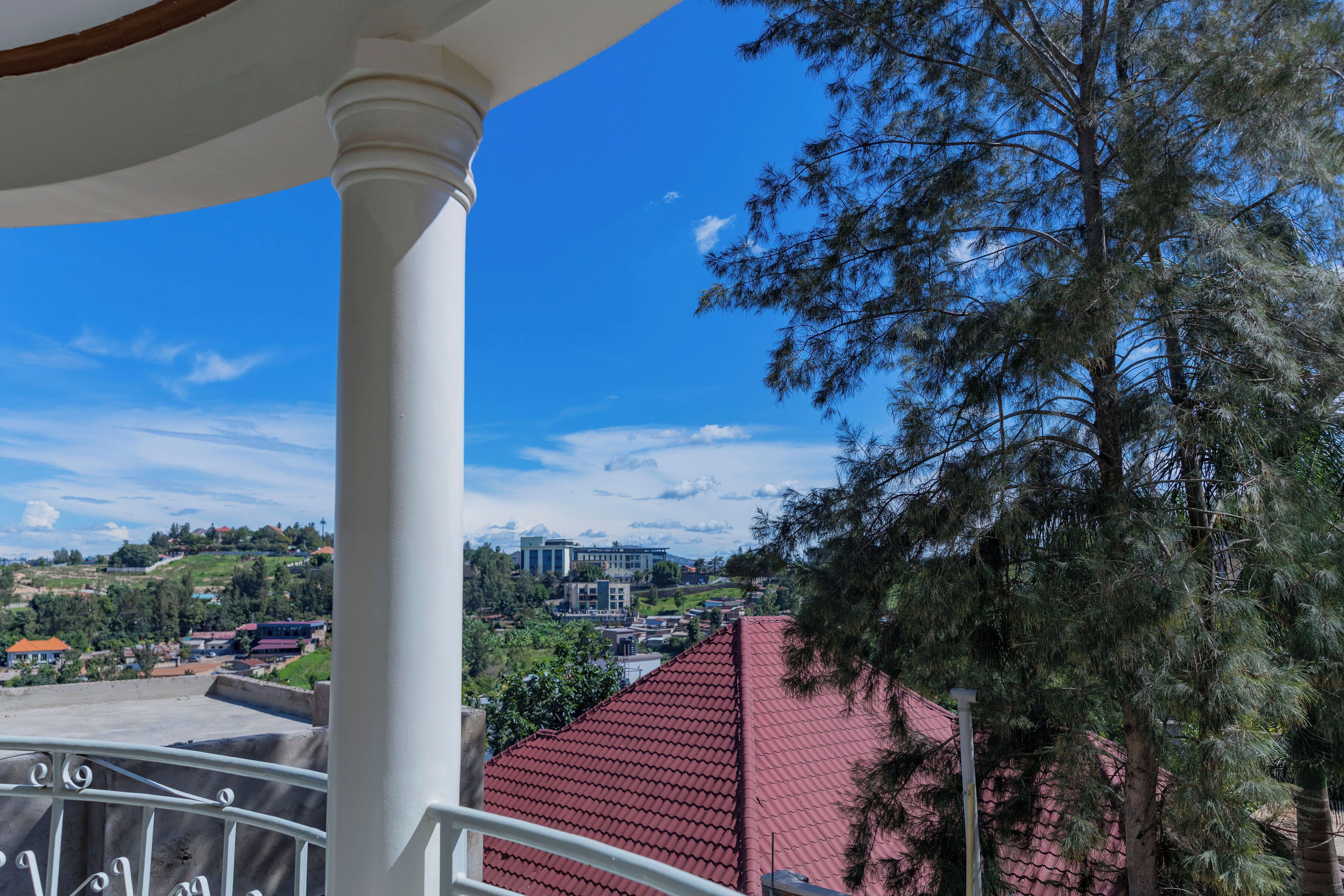 A balcony with a red-tiled roof, white columns, and a view of buildings and trees in the background.
