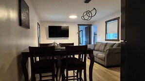 A dining area with a table and chairs, a television mounted on the wall, and a sofa in the adjacent living room.