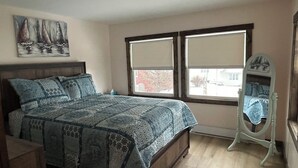 A bedroom with a bed, a window with blinds, a mirror, and a painting on the wall.