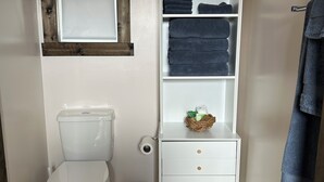A small bathroom with a toilet, a white cabinet with drawers, a shelf with folded towels, and a wooden-framed mirror.