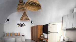 A compact living space with a sofa, ottoman, kitchenette, and two hanging light fixtures. - Imarkellis Pyrgos Traditional House and Belfry Charm with Courtyard in Santorini (Pyrgos Kallistis)