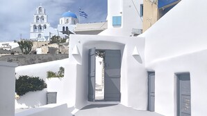 A white-walled building with a doorway leading to a courtyard, a blue-domed church in the background, and a sign reading "A MUSE". - Imarkellis Pyrgos Traditional House and Belfry Charm with Courtyard in Santorini (Pyrgos Kallistis)