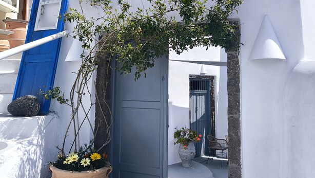 A white building with a blue door, a potted plant, and a bell tower in the background. - Imarkellis Pyrgos Traditional House and Belfry Charm with Courtyard in Santorini (Pyrgos Kallistis)