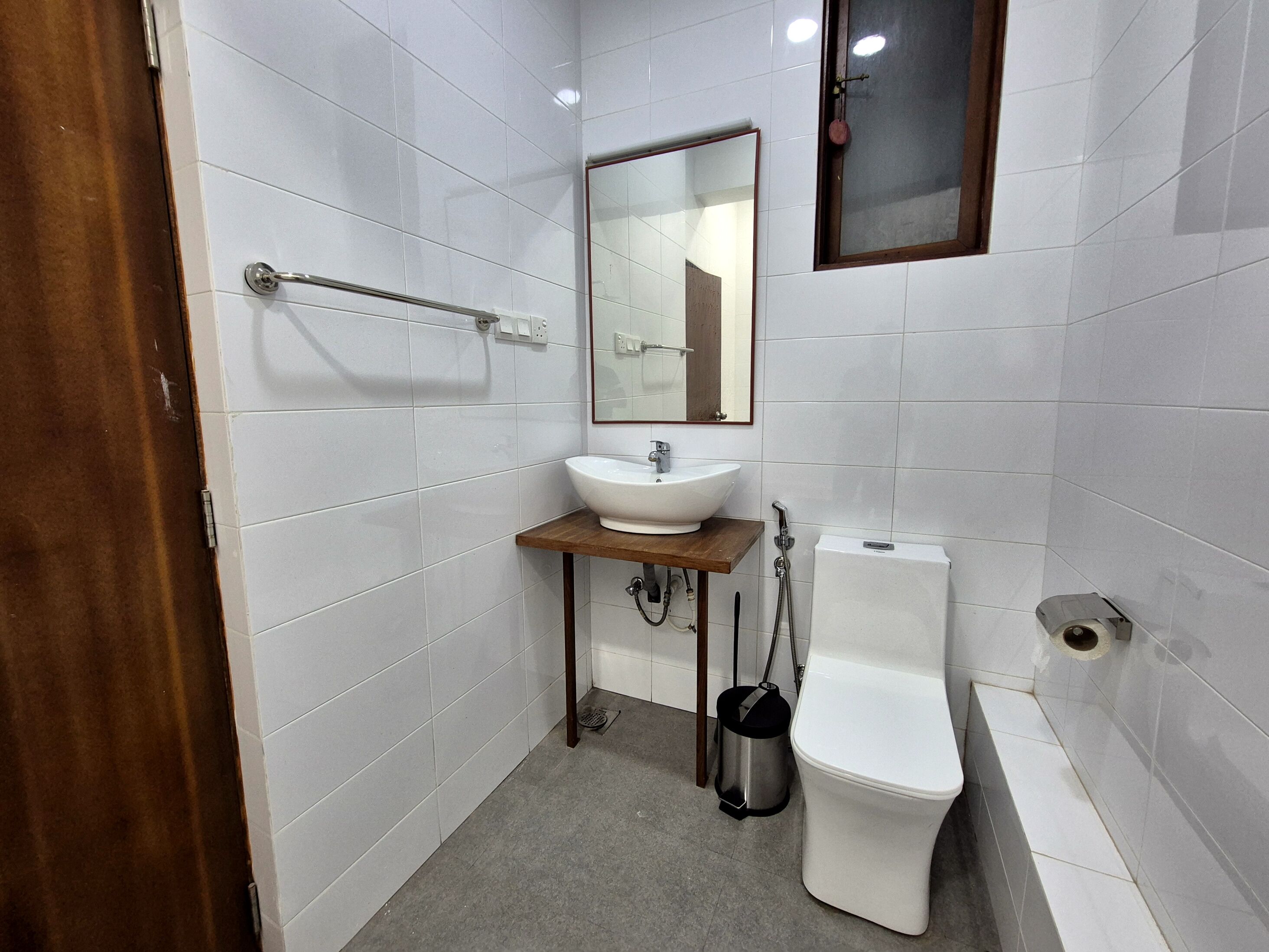 A small, clean bathroom with white tiles, a wooden sink counter, a mirror, a toilet, and a trash can.