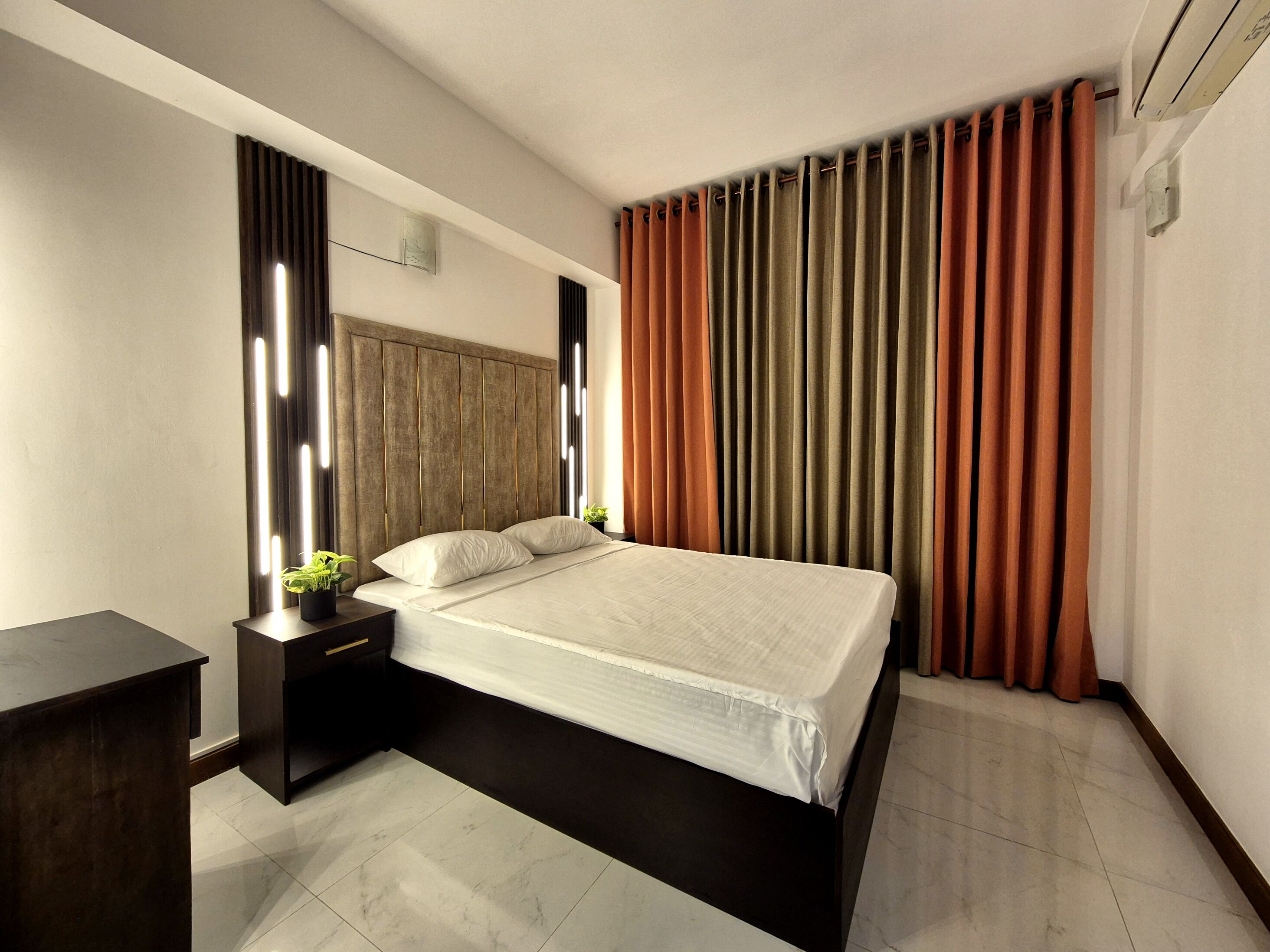 A modern hotel room with a large bed, a nightstand, a small table with a plant, and a large window with curtains.