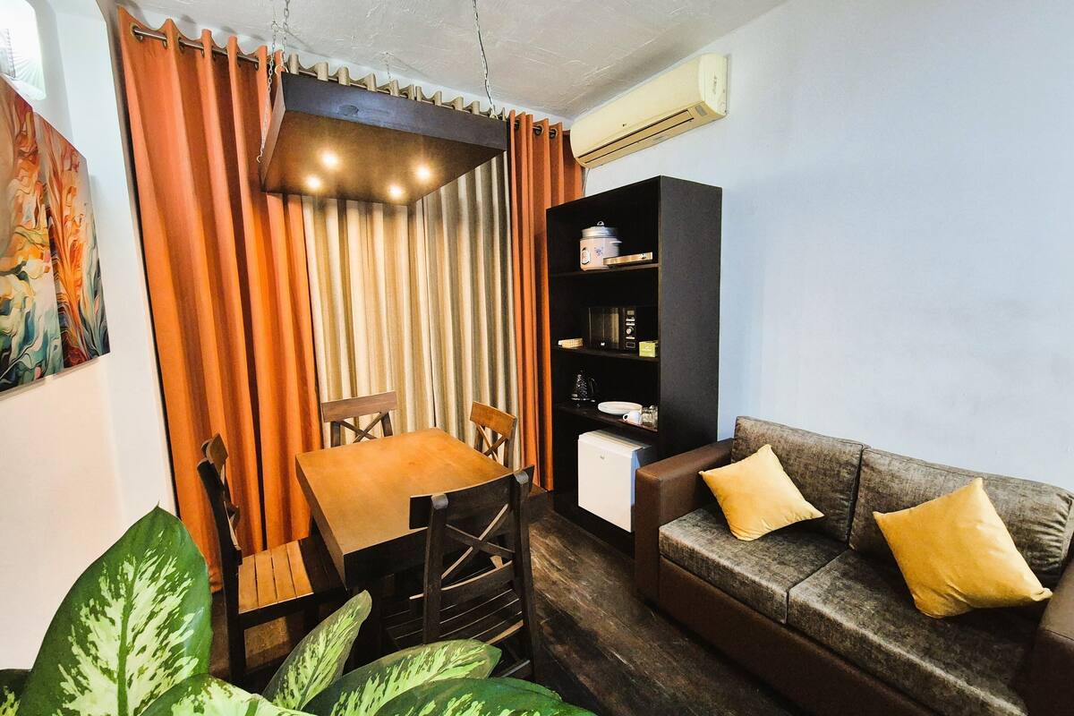 Un espacio habitable compacto con una mesa de comedor y sillas, un sofá con cojines, una estantería y una planta grande en maceta.