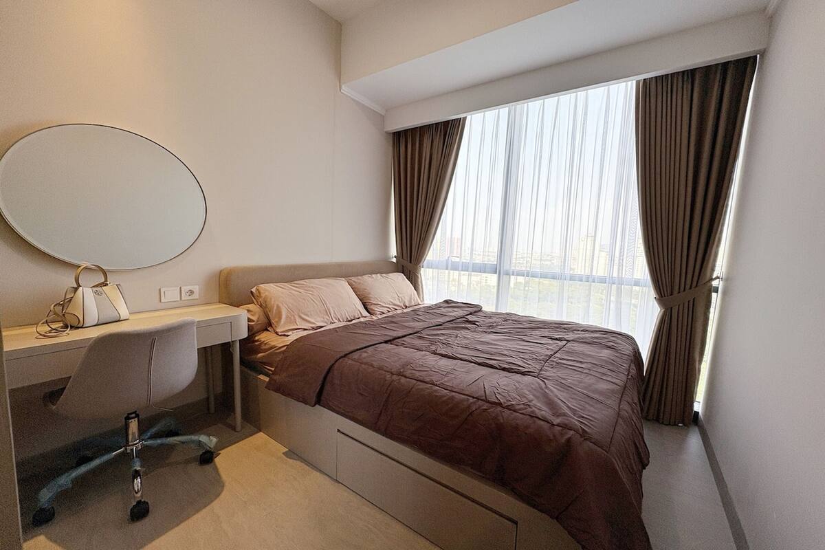 Quarto de hotel com cama, escrivaninha com cadeira, janela grande com cortinas e um espelho redondo na parede.