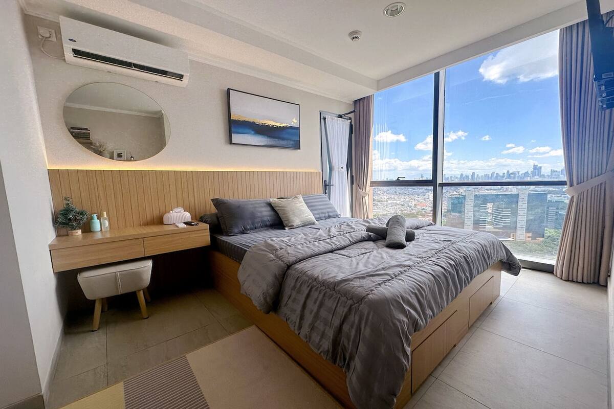 Quarto moderno com uma cama grande, uma escrivaninha, um espelho e vista para a cidade.