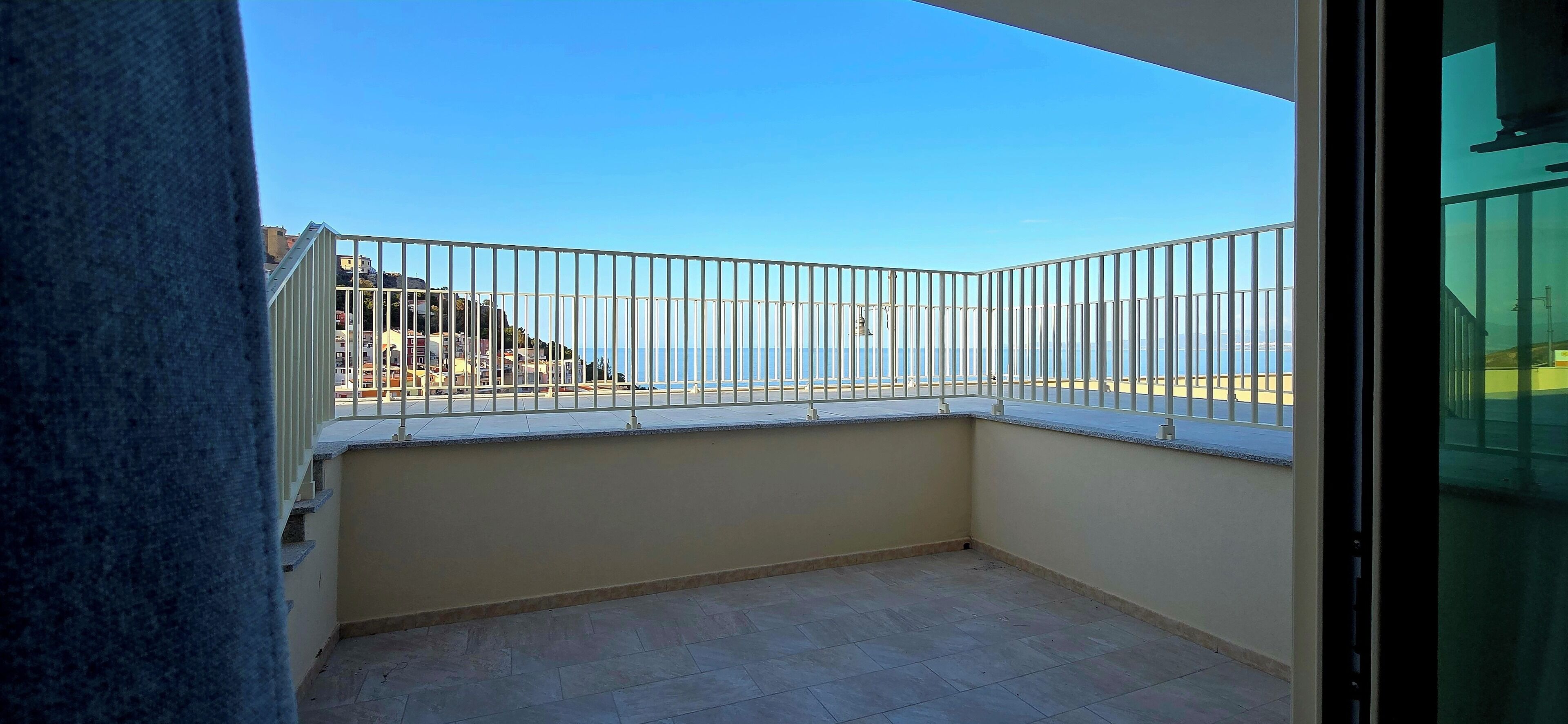 Uma varanda com guarda-corpo de metal, vista para o mar e um edifício ao fundo.