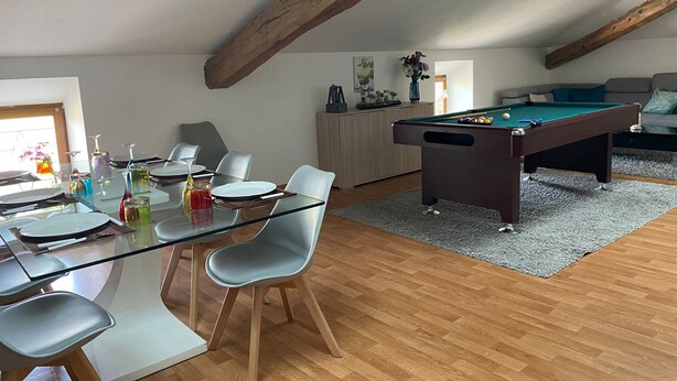 A dining area with a glass table and chairs, a pool table, and a sofa in a room with wooden beams and a gray carpet. - Billiards Lounge loft with Billiards & Netflix by BoookingFuté (Socourt)