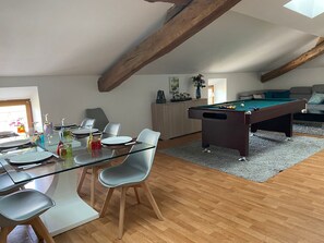 A dining area with a glass table and chairs, a pool table, and a sofa in a room with wooden beams and a gray carpet.