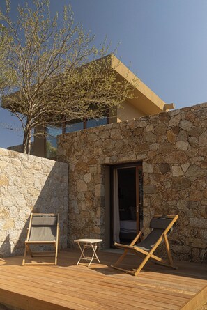 A wooden deck with two lounge chairs and a small table, a stone wall, and a door leading to a room.
