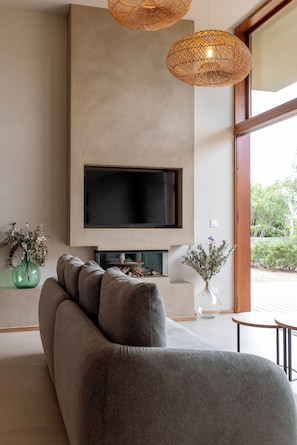 A modern living room with a large flat-screen TV, a grey sofa, wicker pendant lights, and a view of greenery outside.