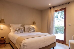 Ein Schlafzimmer mit einem Bett, zwei Nachttischlampen, einem Fenster mit Vorhängen und Blick ins Grüne.