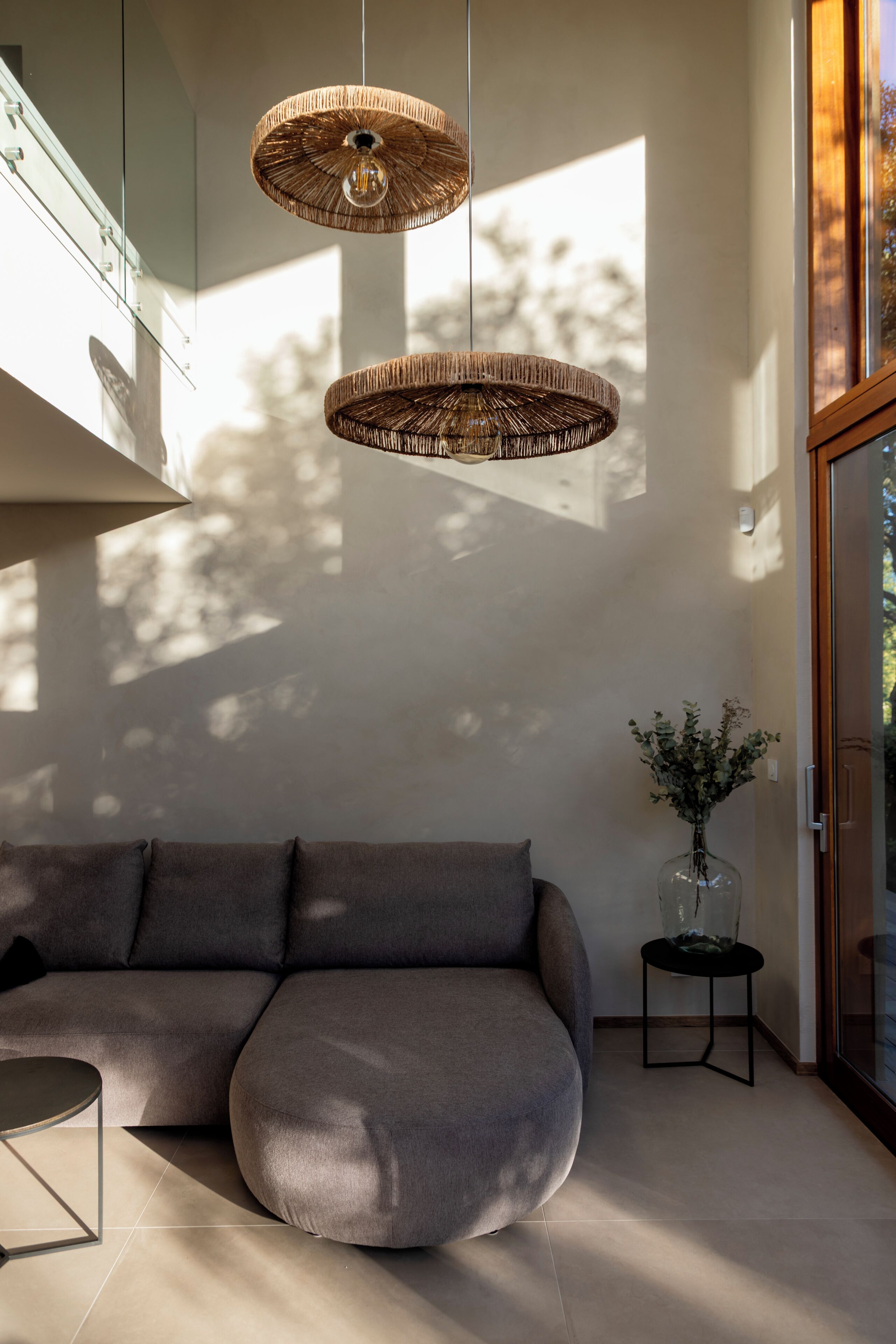 A modern living room with a sectional sofa, two woven pendant lights, a small side table with a vase, and a glass door leading to an outdoor area.
