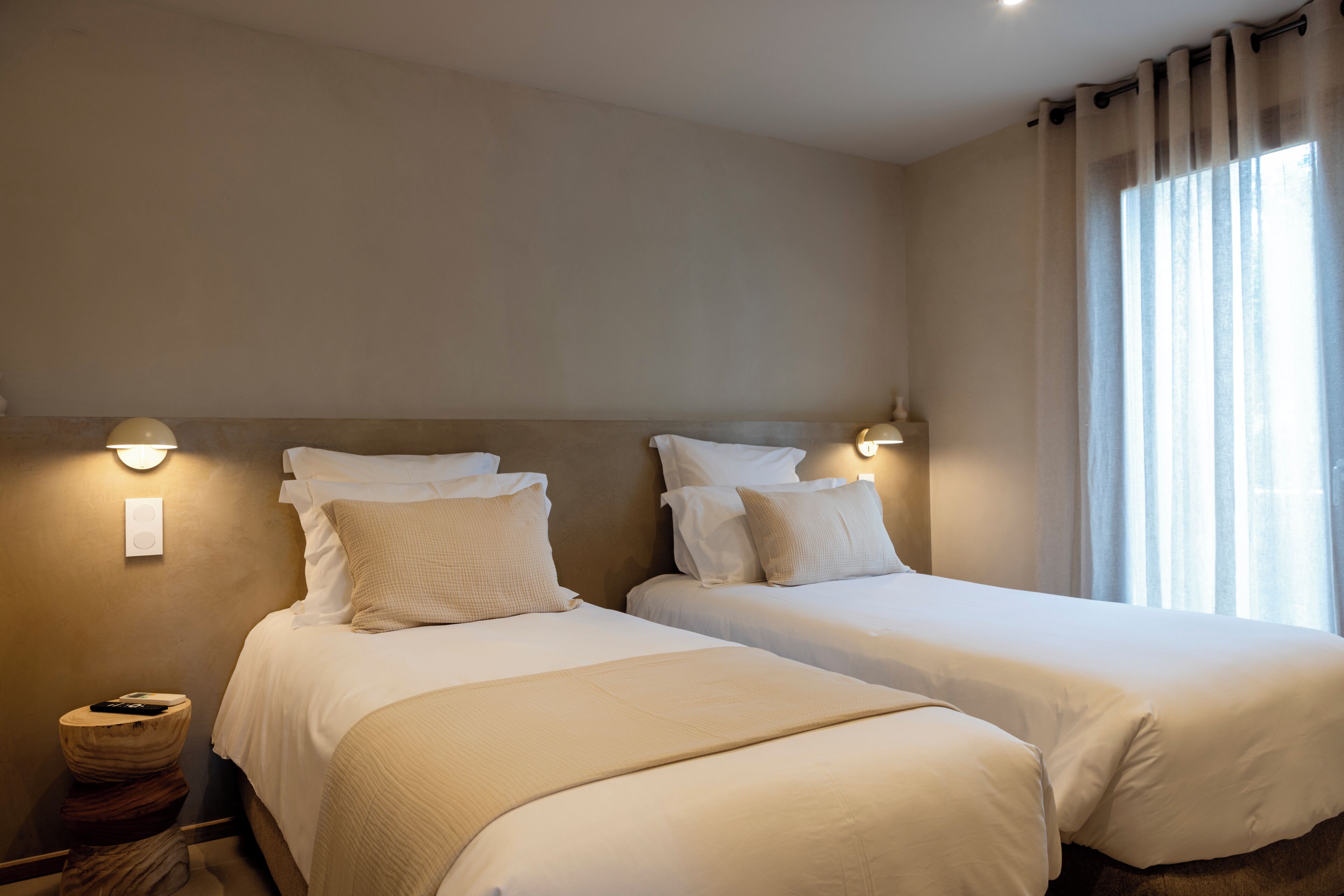 A hotel room with two beds, a bedside table, a lamp, and a window with curtains.