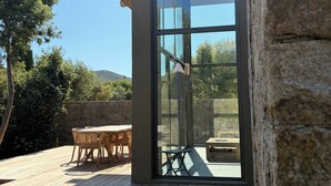 A modern outdoor patio with a wooden deck, a glass door leading to an interior space, and a stone wall. - Villa with swimming pool, near Lozari beach – Corsica (BELGODÈRE)