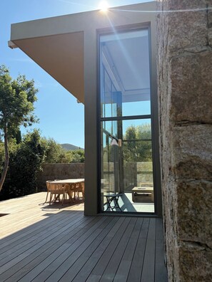 A modern outdoor patio with a wooden deck, a glass door leading to an interior space, and a stone wall.
