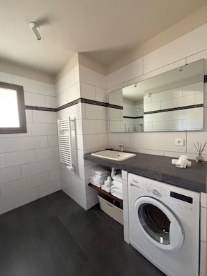 A modern bathroom with a built-in sink, a large mirror, a towel rack, a washing machine, and a window.