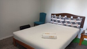 Una cama bien hecha con un colchón blanco y un cojín estampado, una mesita de noche de madera con un control remoto y una silla azul turquesa frente a una pared lisa.