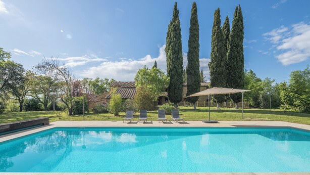 Ein rechteckiger Swimmingpool mit klarem, blauem Wasser, umgeben von einer gefliesten Liegefläche. Vier Liegestühle sind entlang des Poolrandes aufgereiht, und ein Sonnenschirm spendet Schatten. Im Hintergrund sind hohe, schlanke Bäume zu sehen, und hinter den Bäumen befindet sich ein Gebäude mit einem gefliesten Dach.