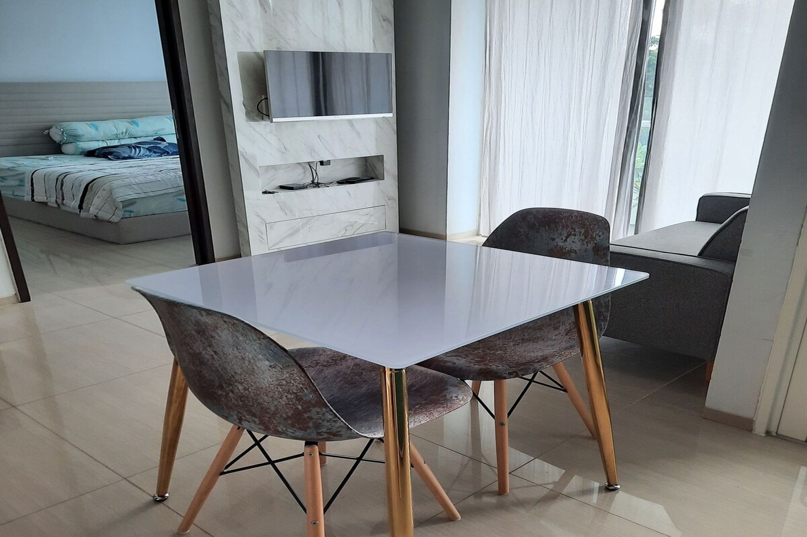 A modern living room with a glass-top dining table, two chairs, a sofa, a TV mounted on a marble wall, and a large window with curtains.