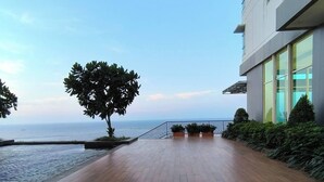A wooden deck with a view of the sea, a modern building with large windows, and a tree in the center.