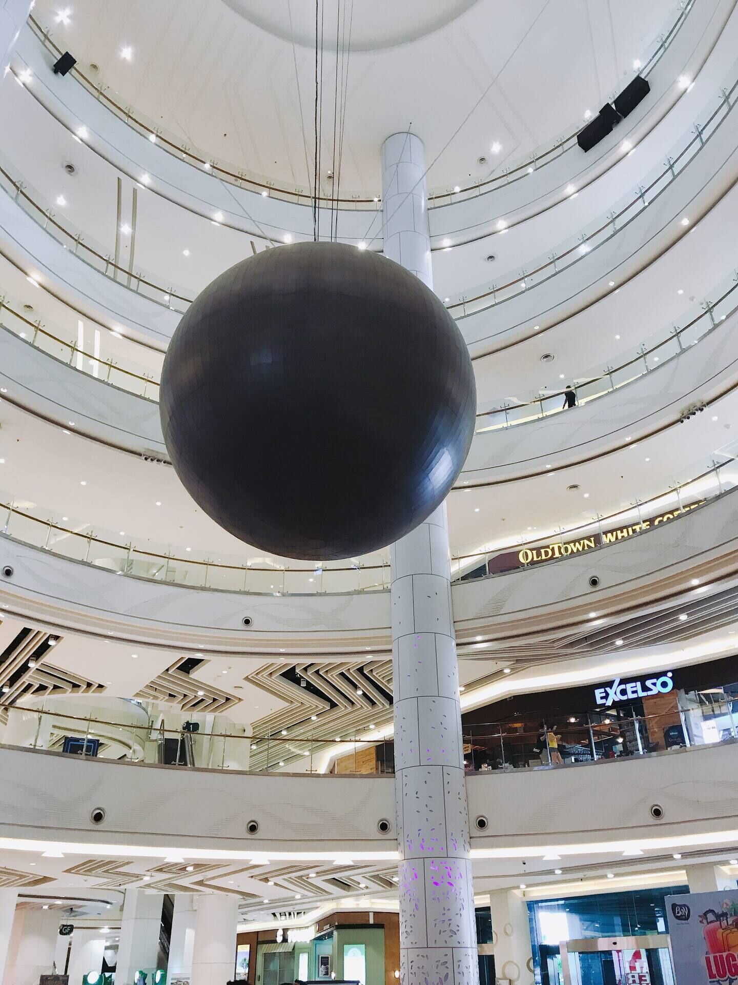 Een meerlaags winkelcentrum met een grote, bolvormige sculptuur aan het plafond, meerdere verdiepingen met glazen hekwerk en diverse winkelpanden, waaronder OldTown White Coffee en Excelso.