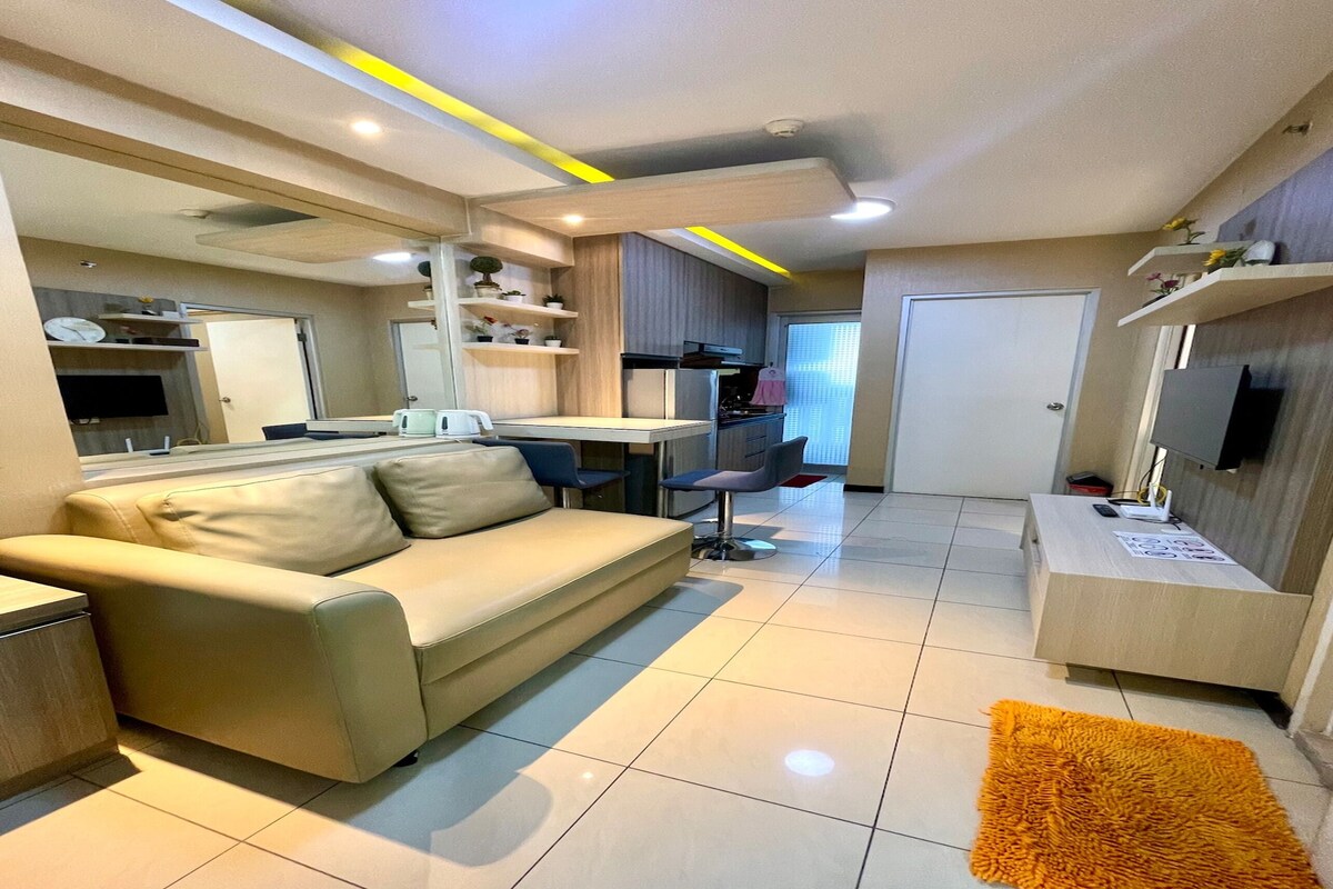 Una sala de estar moderna con un sofá, una mesa de centro y un televisor. Cuenta con una zona de cocina con armarios y un refrigerador.