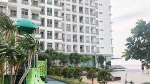 A multi-story residential building with balconies, a playground with green equipment, and a paved area with trees and shrubs. - Sky Heaven Greenbay in North Jakarta (Jakarta)