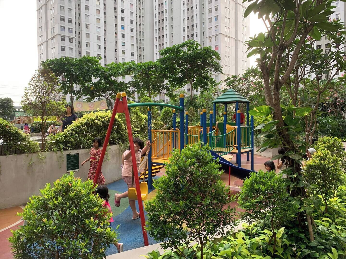 Un parque infantil con estructuras para escalar y un tobogán, rodeado de vegetación y edificios altos al fondo.