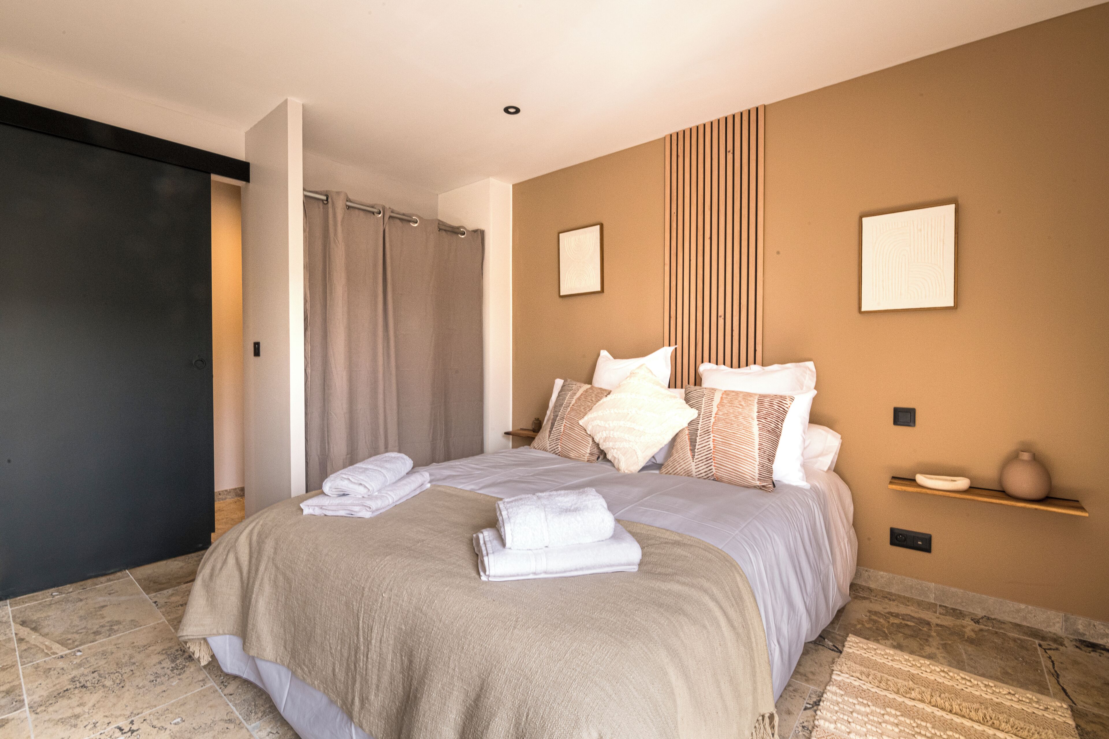 Une chambre moderne avec un grand lit, une porte sombre, un rideau de douche et une étagère murale ornée d’un vase.