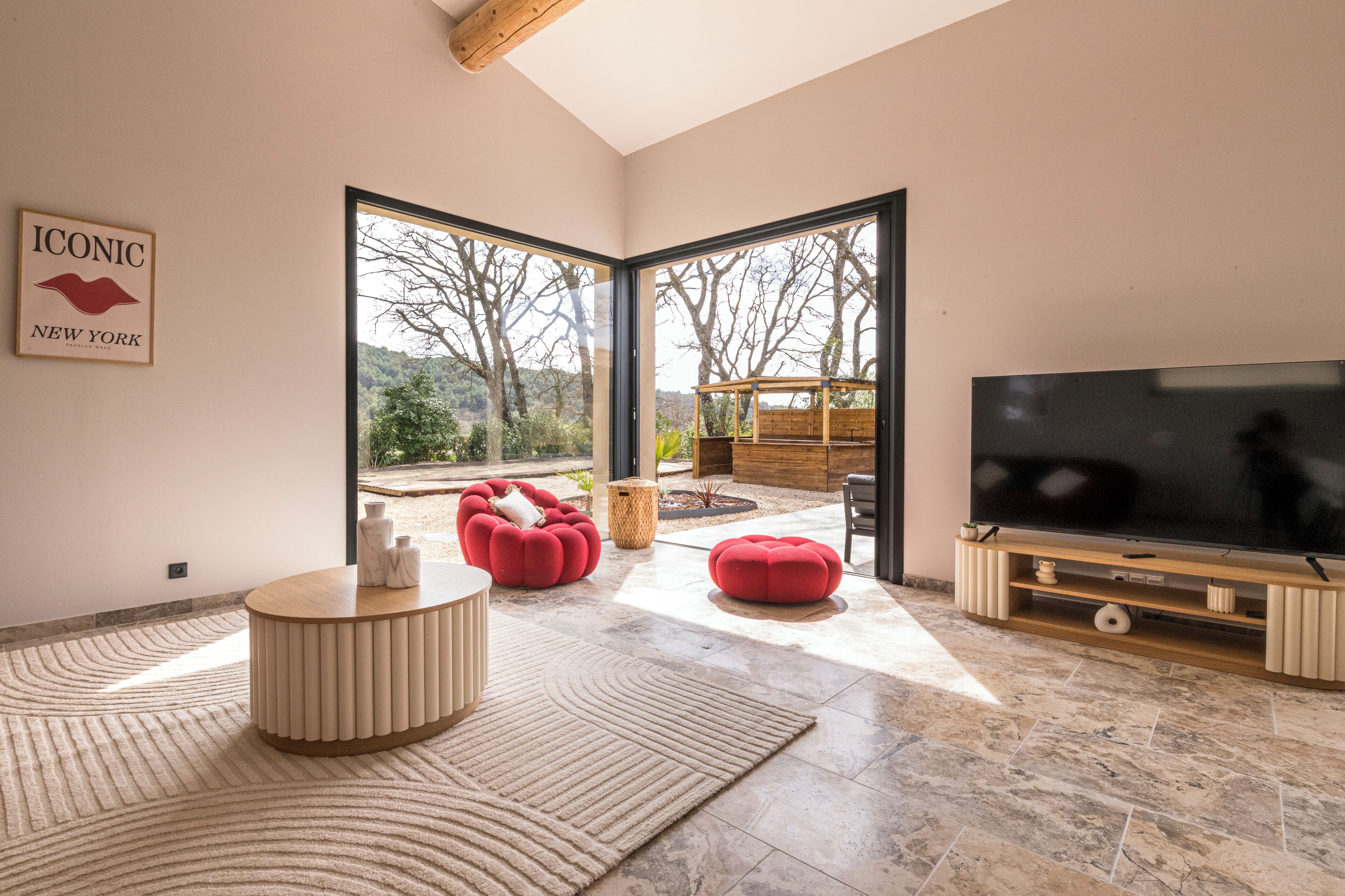 A modern living room with a large TV, red ottomans, a wooden coffee table, and a view of the outdoors.