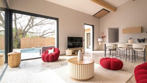 A modern living room with a large TV, red seating, a wooden coffee table, and a kitchen area with bar stools. - Villa with pool, outdoor bar and balinese bed (Cadenet)