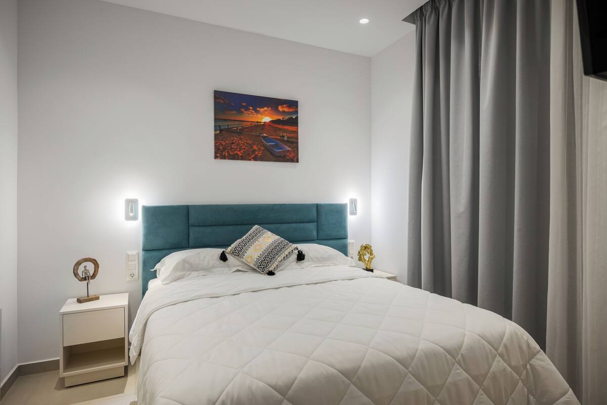 Un dormitori amb un llit, una tauleta de nit, un quadre penjat a la paret i una finestra amb cortines.