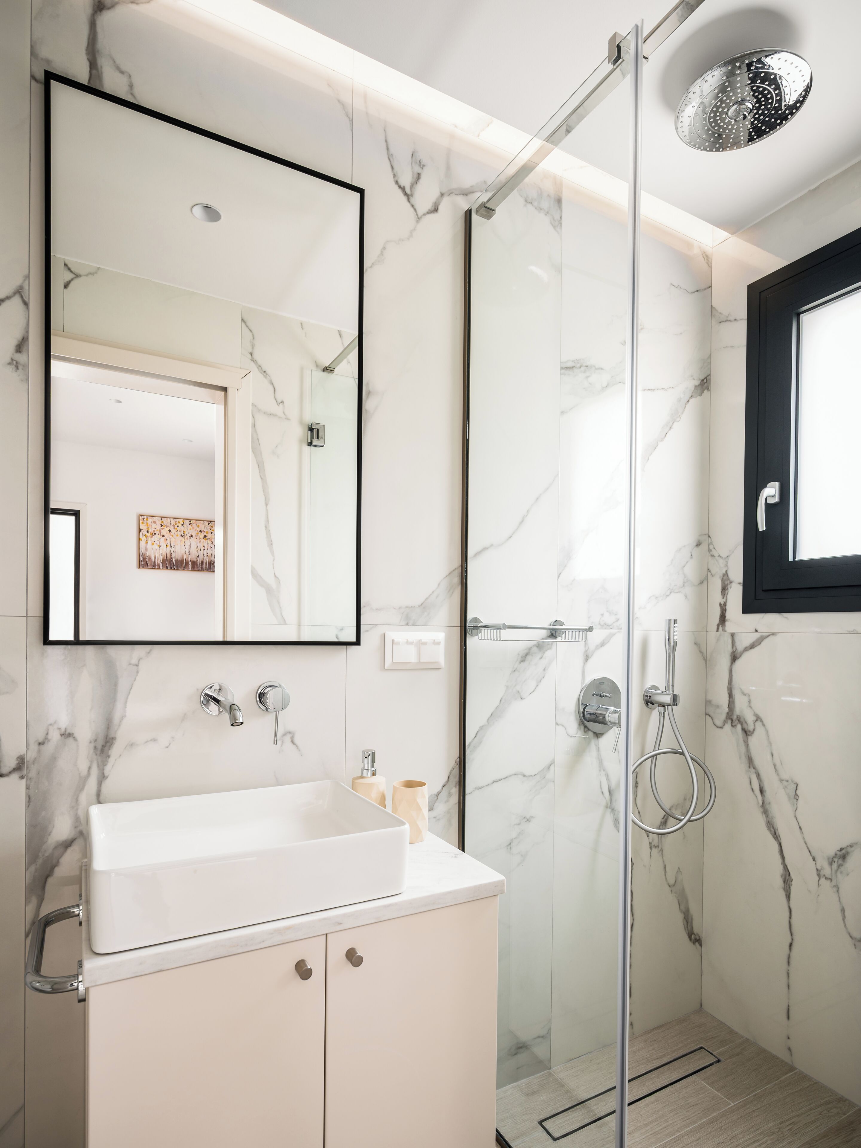 Une salle de bain moderne avec un mur en marbre, une douche à parois de verre, un lavabo blanc et un grand miroir.