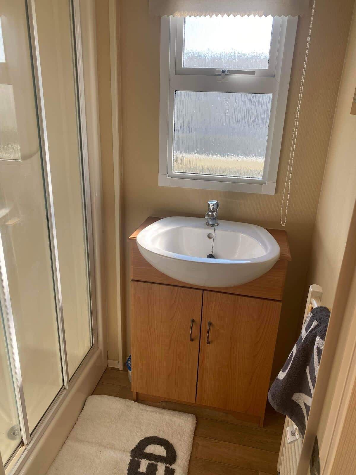 A bathroom with a white sink, wooden cabinets, a window, and a towel on the floor.