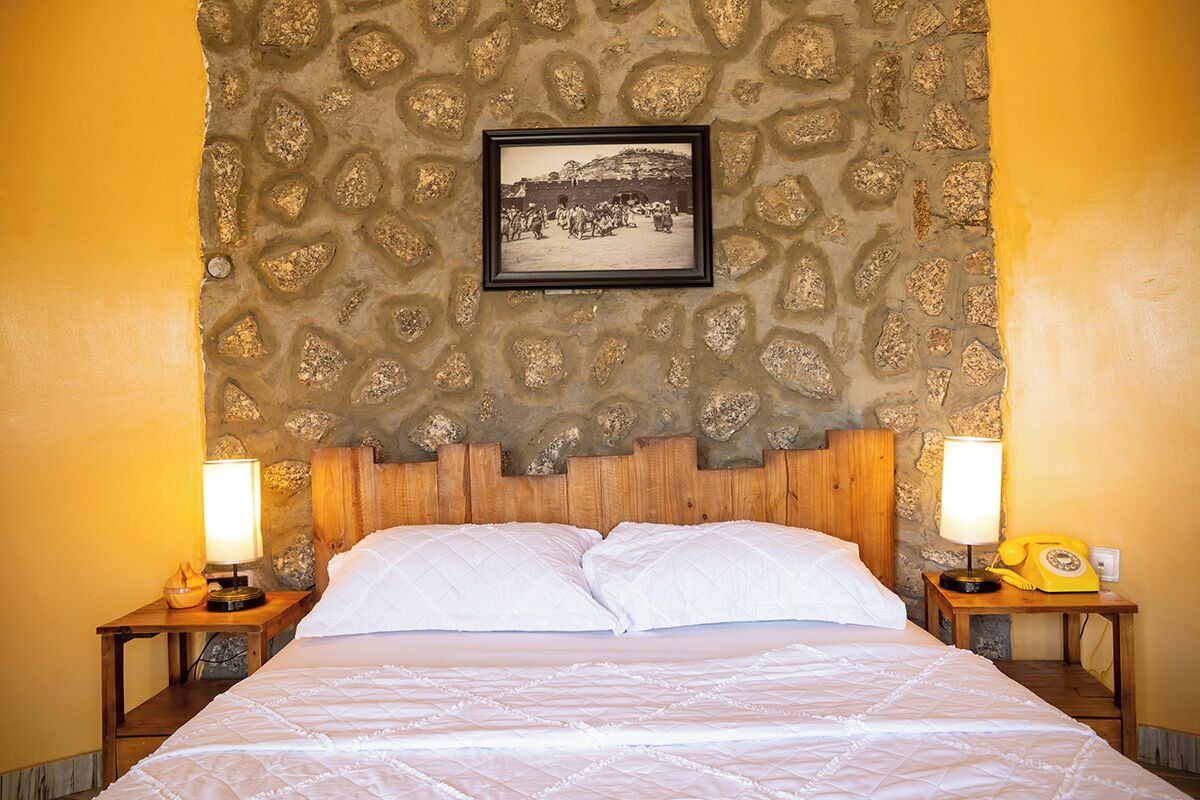 Una cama con ropa blanca, una pared de piedra con un cuadro enmarcado y dos lámparas de noche sobre mesitas de madera.