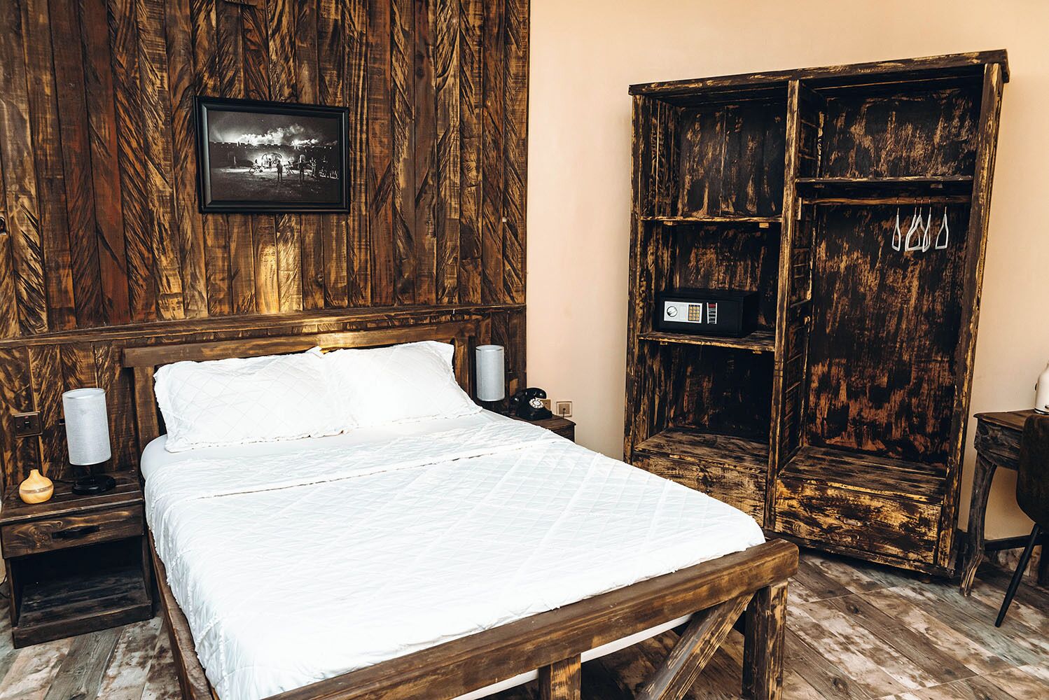 A room with a wooden bed, a wooden wardrobe, a framed picture on the wall, and a bedside table with a lamp.
