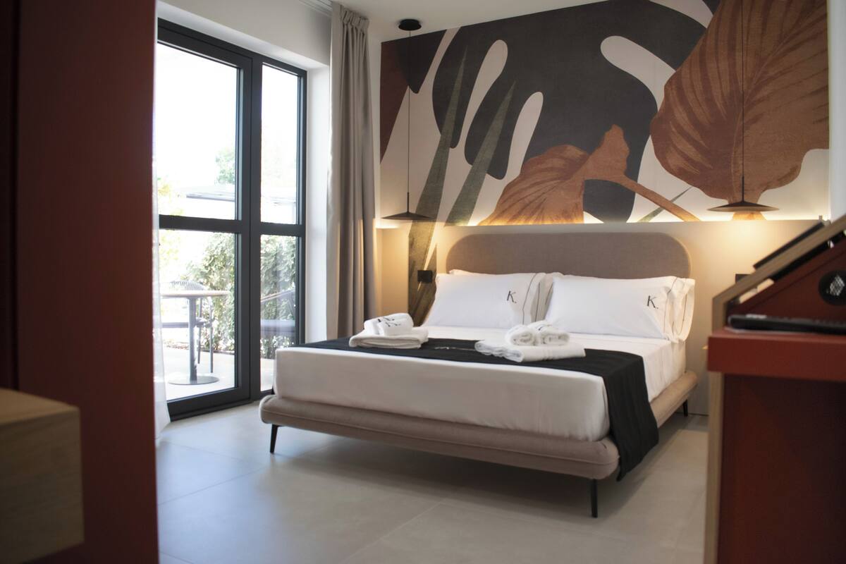 Une chambre d’hôtel moderne avec un grand lit, une vue à travers une porte vitrée et une grande œuvre d’art abstraite accrochée au mur.