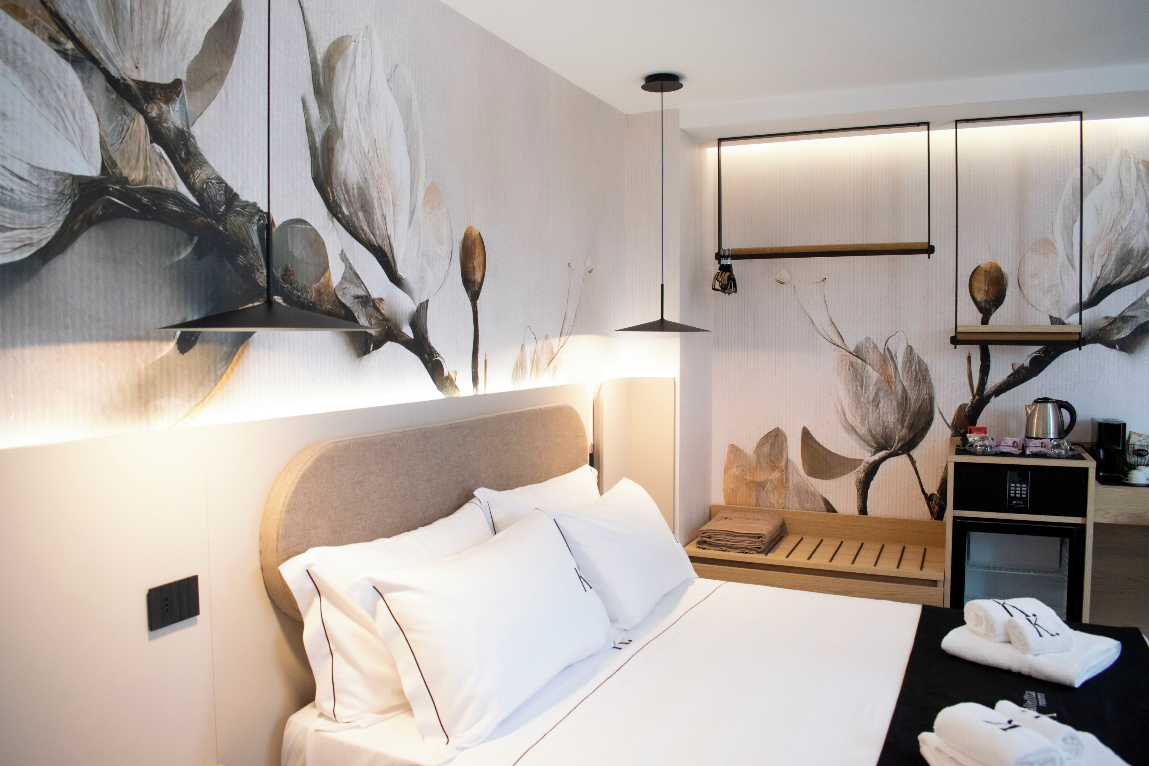 A hotel room with a bed, a nightstand, a wall-mounted shelf with a kettle, and a large abstract painting.