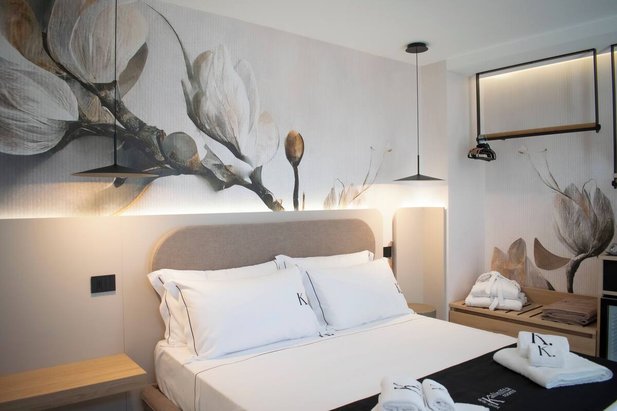Une chambre d’hôtel avec un lit, une table de chevet, une étagère fixée au mur où sont posées des serviettes, et un grand tableau représentant des fleurs accroché au mur.