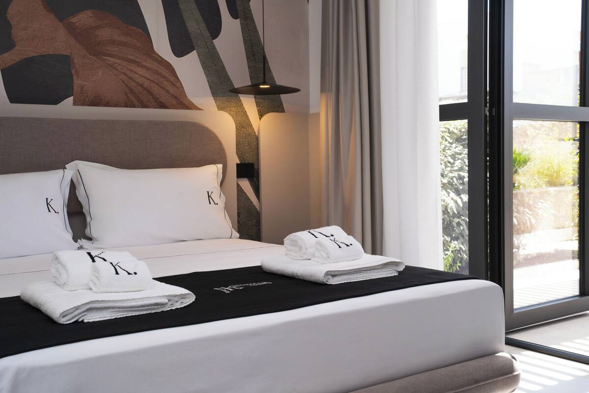 Une chambre d’hôtel moderne avec un grand lit, deux oreillers ornés d’un logo, une tête de lit noire, une table de chevet noire et une fenêtre donnant sur un paysage verdoyant.