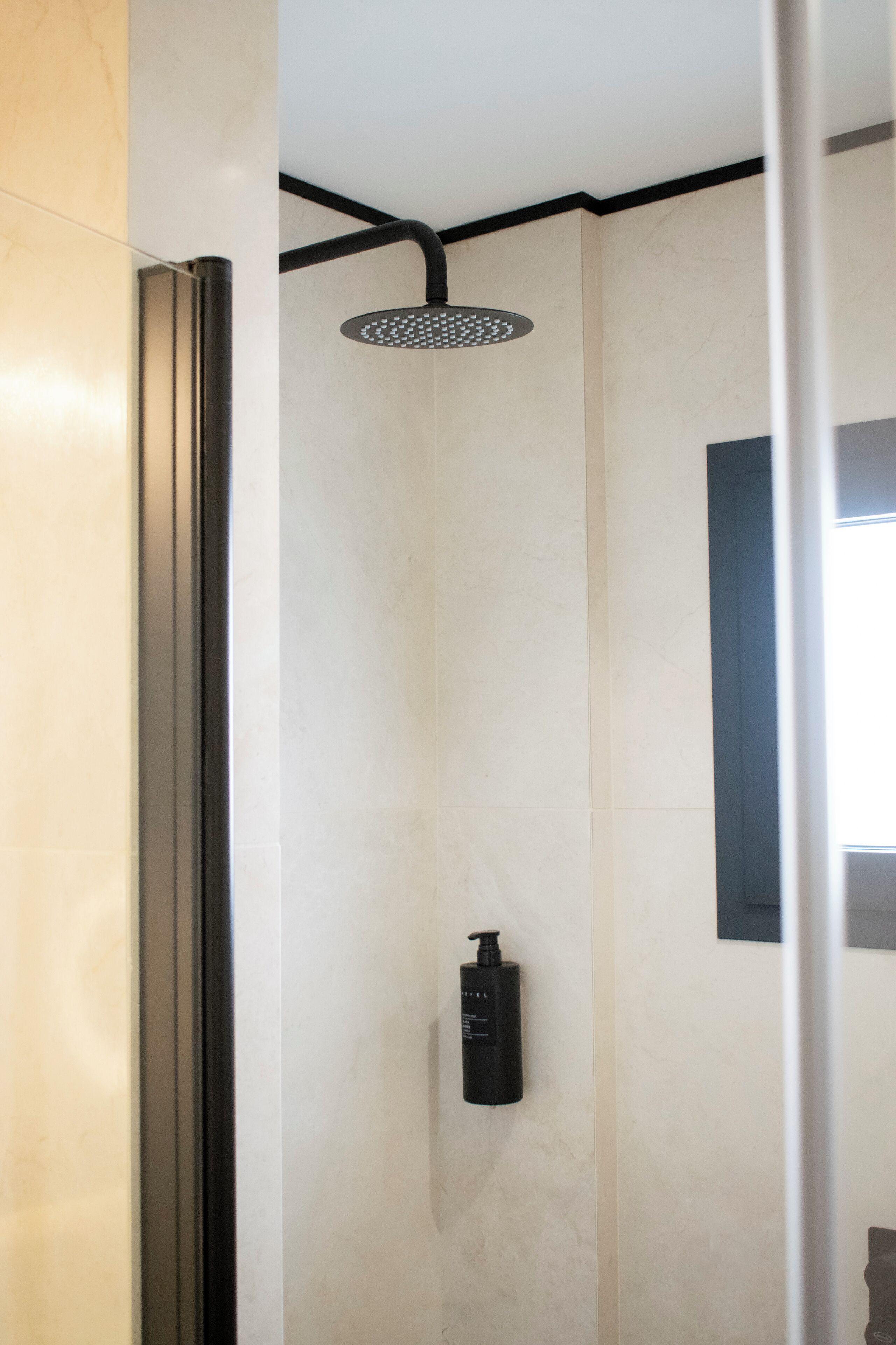 A modern shower with a handheld showerhead, a black soap dispenser, and a small window.