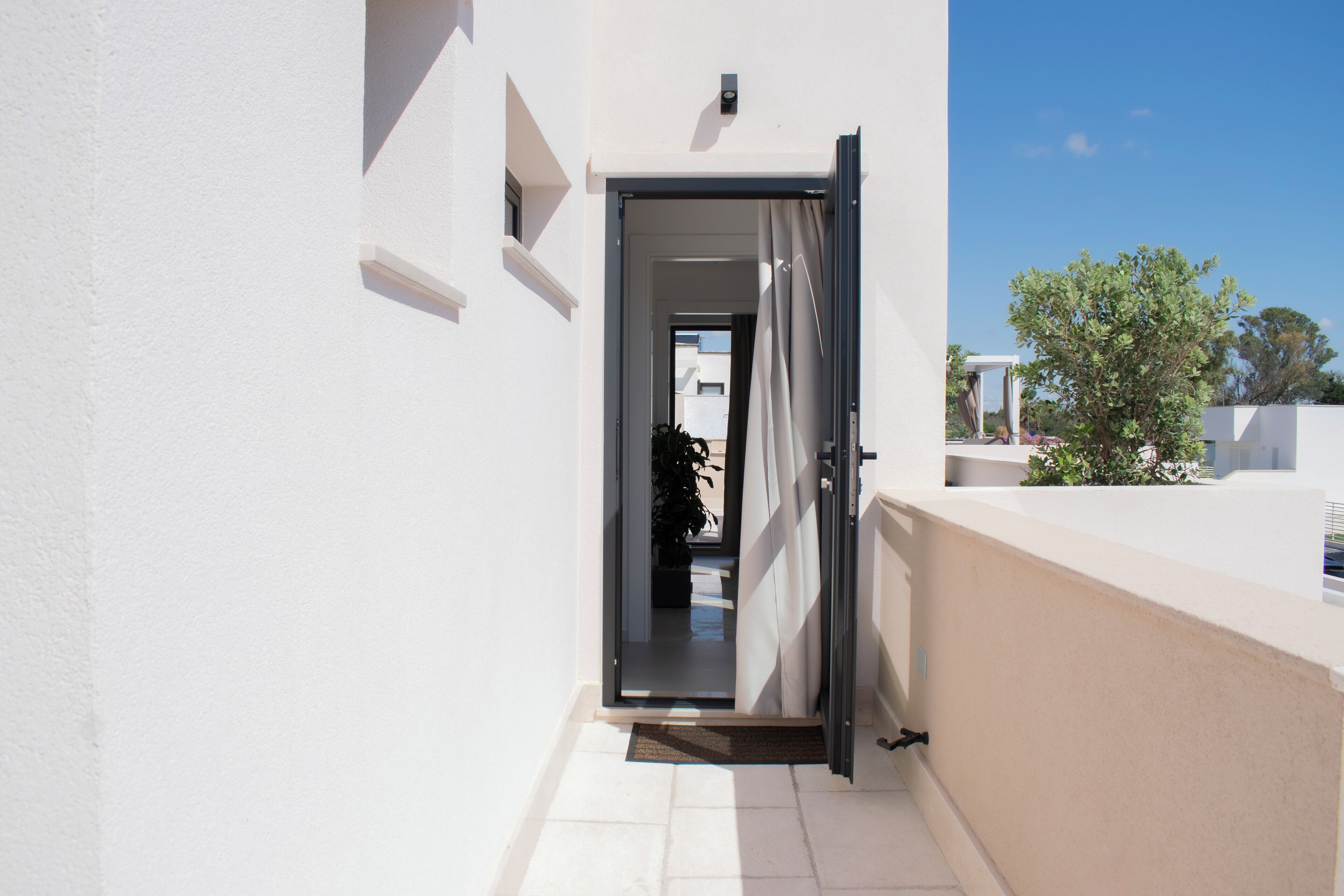 Une porte ouverte donnant sur un balcon, d’où l’on peut admirer un arbre et un bâtiment en arrière-plan.